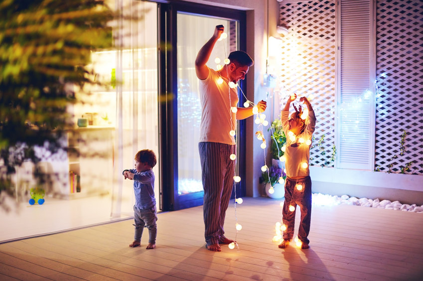 homme avec 2 enfants en train de décorer sur la terrasse d'une maison des guirlandes lumineuses